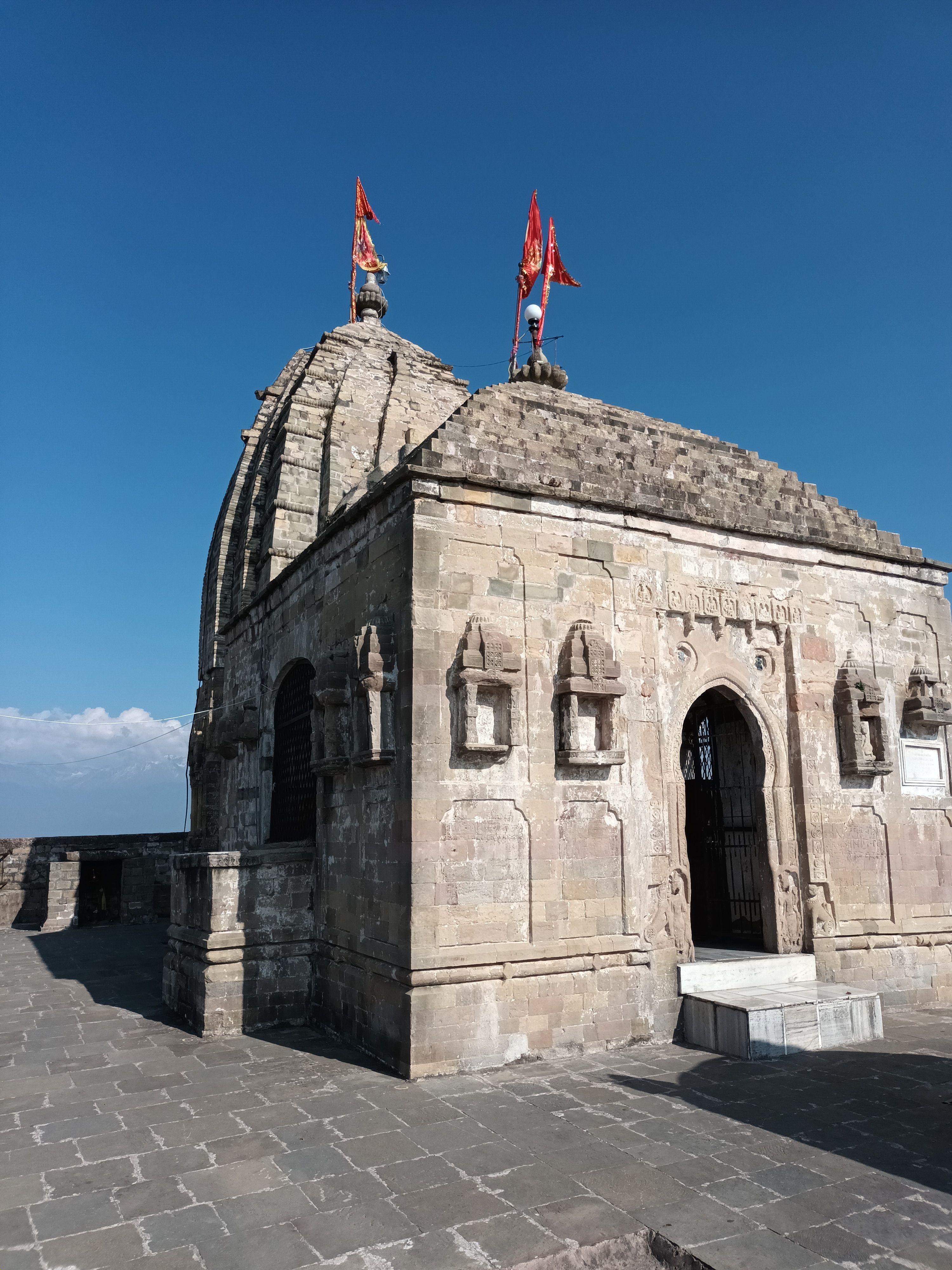 An ancient stone temple with red flags on a sunny day in Himachal Pradesh, India.