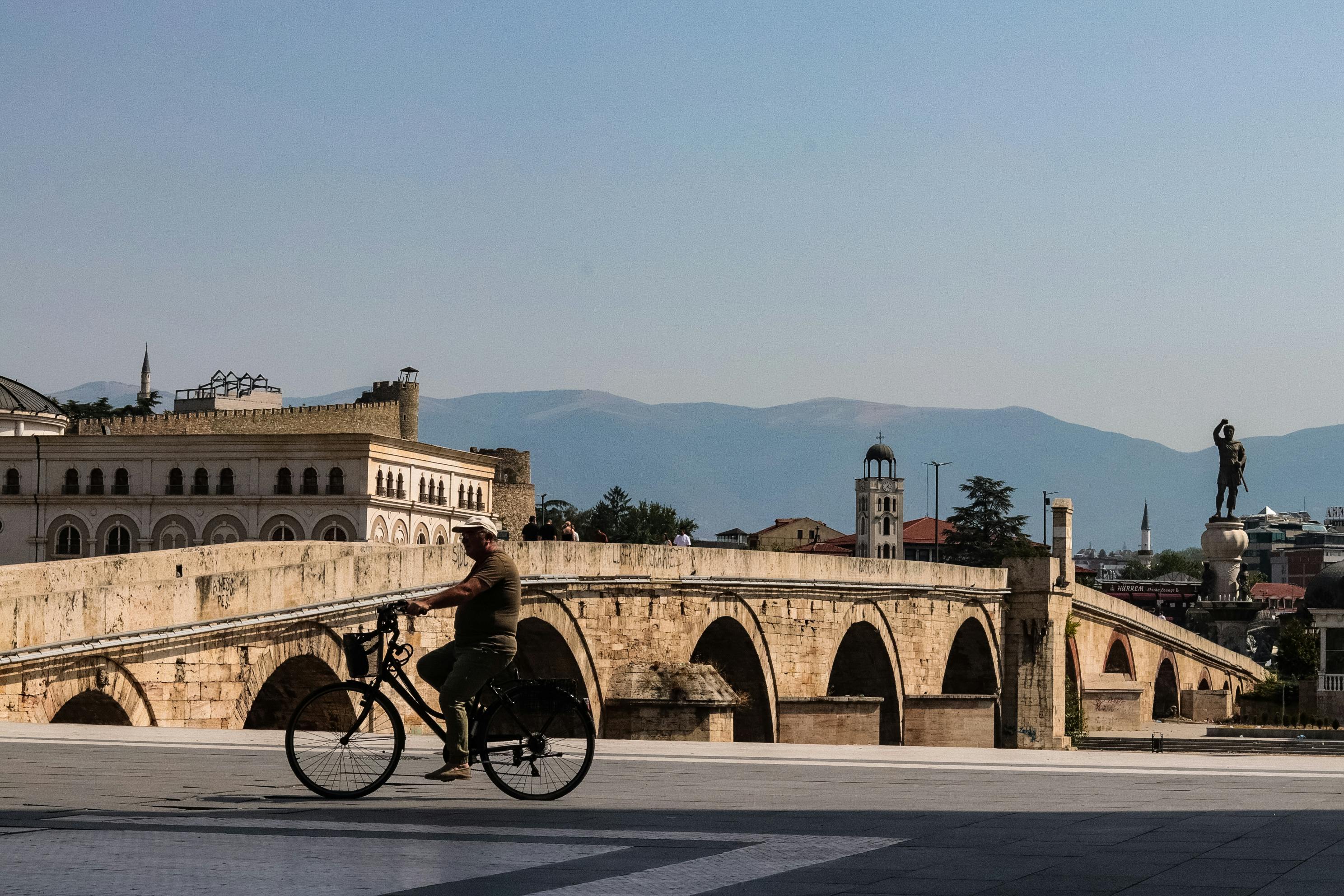 A cyclist rides across the historic Stone Bridge in Skopje, Macedonia against a mountain backdrop.