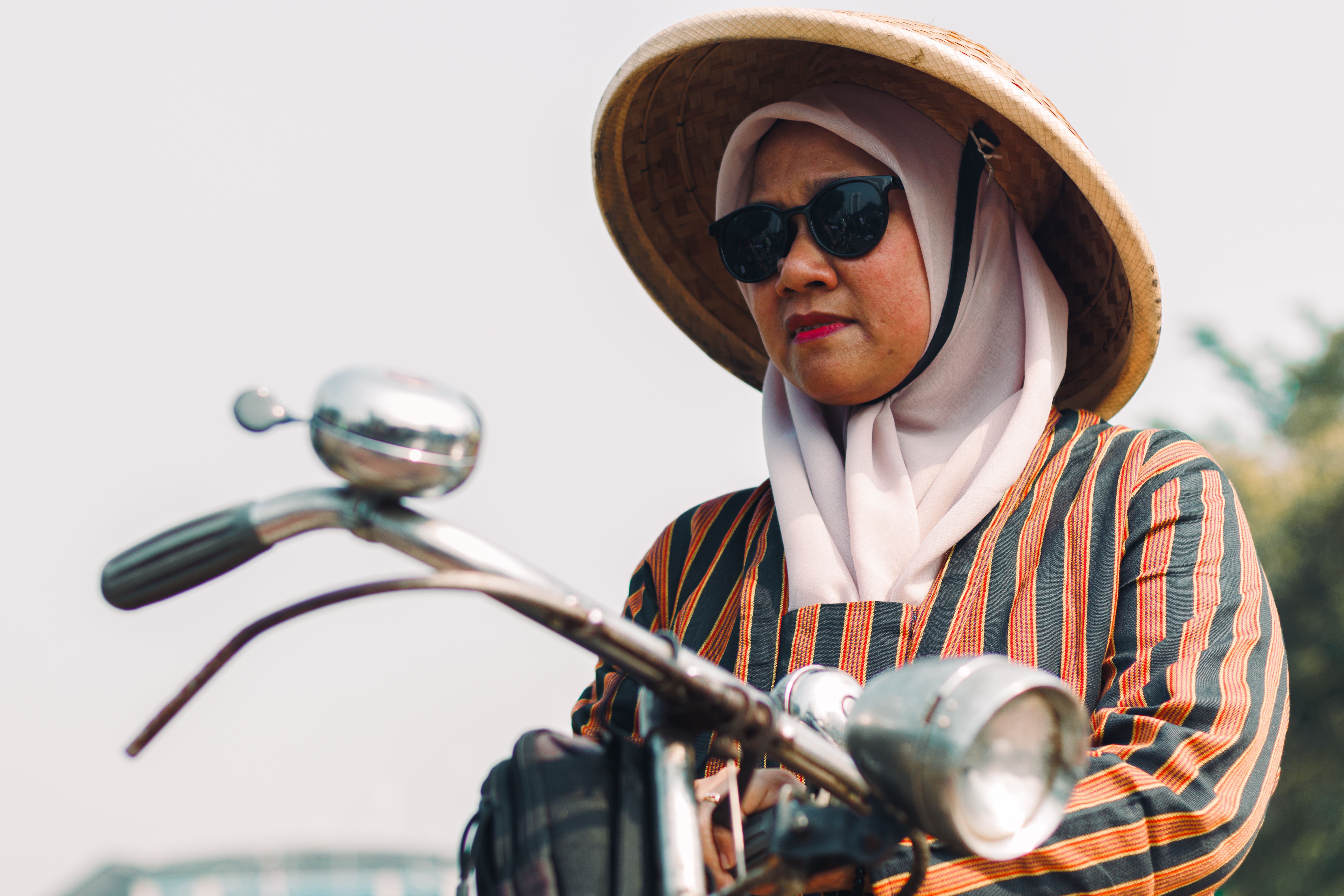 Stylish woman wearing sunglasses and hijab rides a vintage bicycle in a sunny setting, embracing a unique cultural blend.