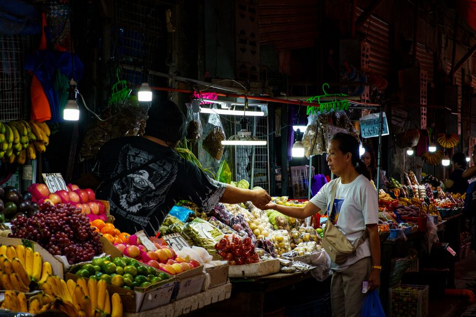 A bustling night market scene with a vendor and a customer exchanging goods surrounded by colorful fruits.