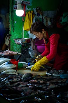 A woman in yellow gloves skillfully prepares fish at an indoor market stall, showcasing daily market life.