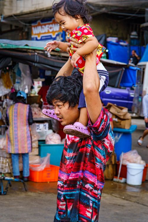 Free Father carrying daughter on shoulders through a vibrant street market with stalls and goods around. Stock Photo