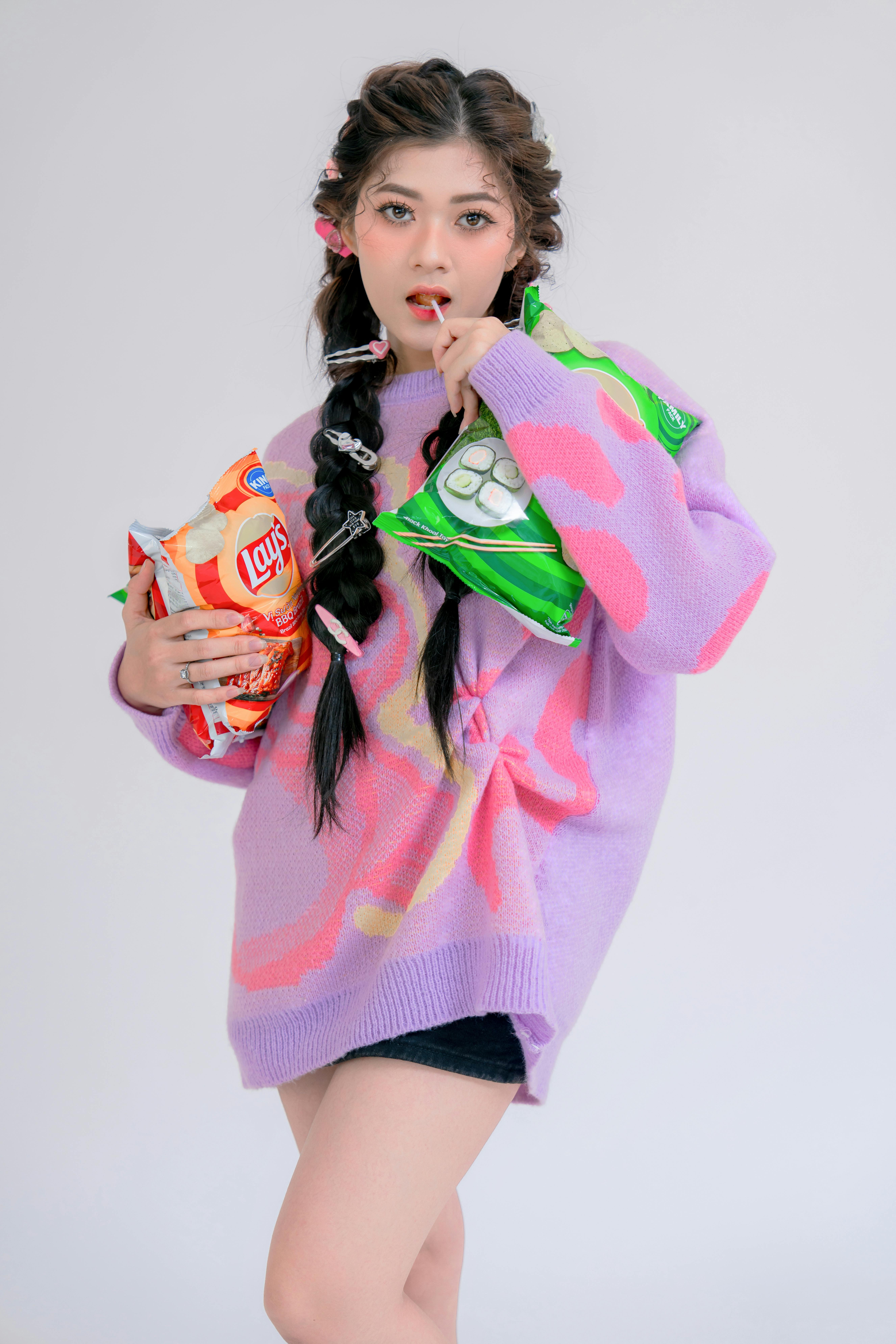 A young woman in a colorful sweater holding snack bags in a studio setting.