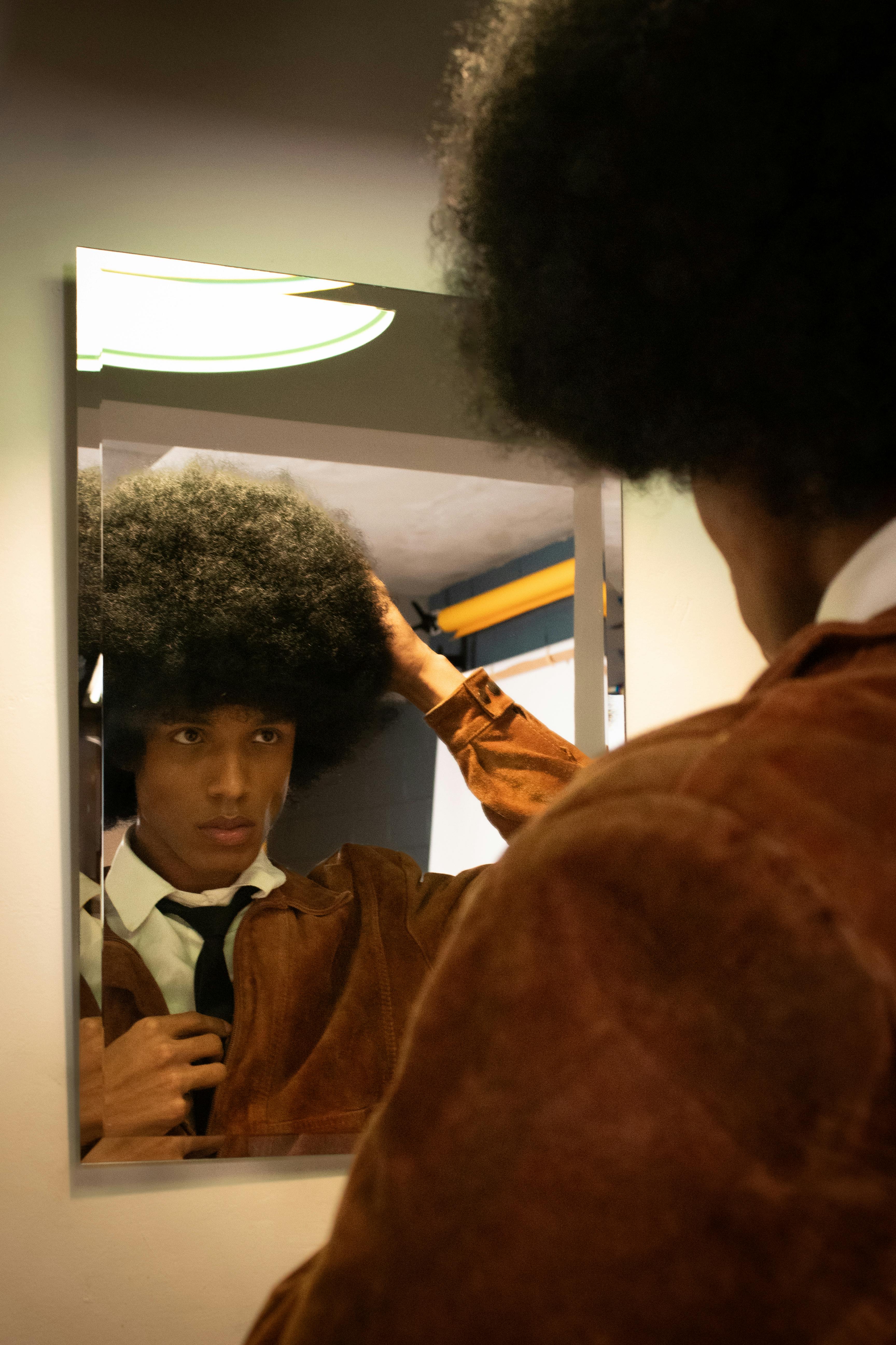 A young adult adjusts their hairstyle in a mirror, wearing a vintage jacket and tie.