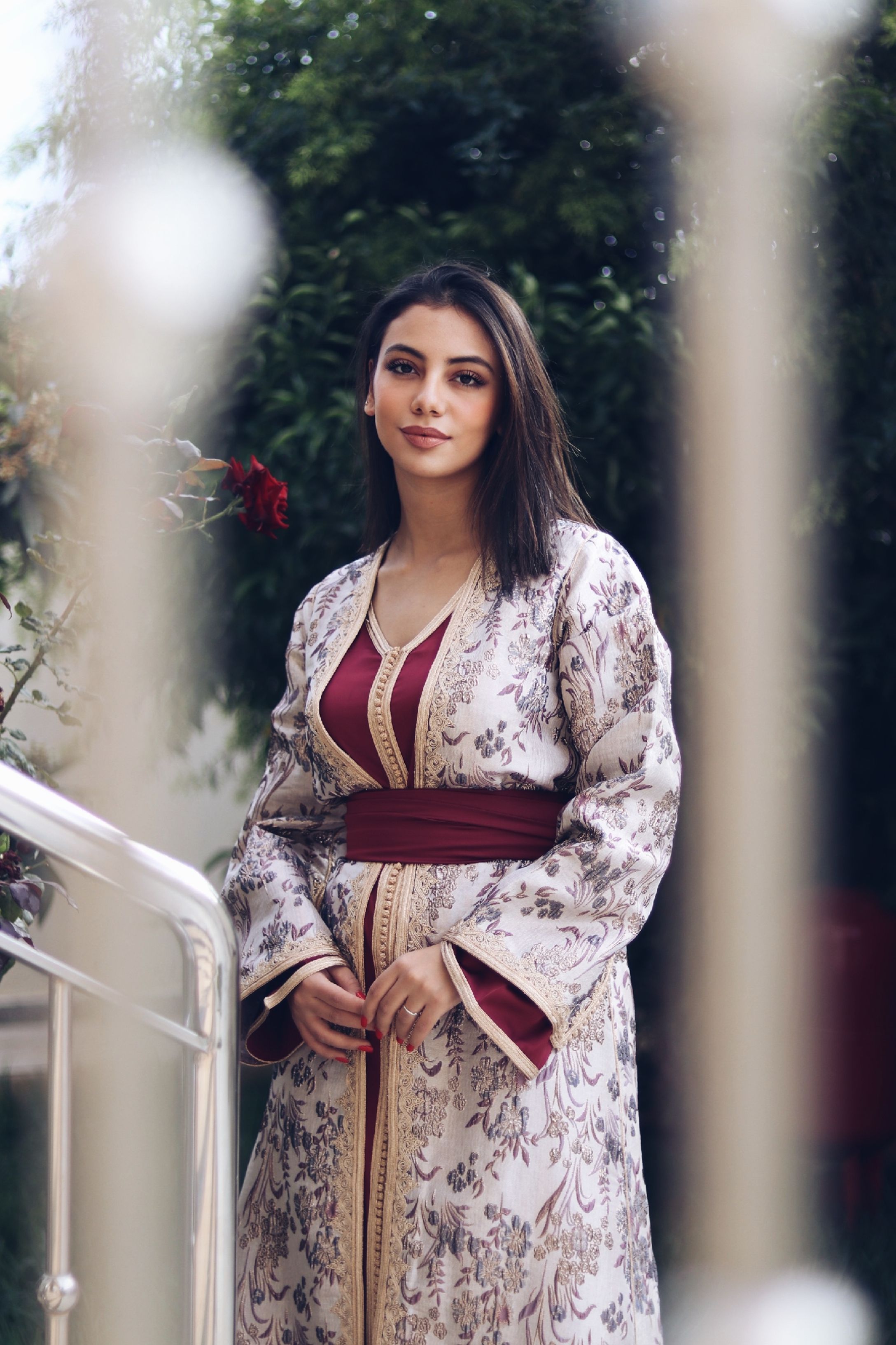 Woman in floral dress poses outdoors, wearing elegant traditional attire.