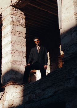 Man in a suit and sunglasses poses dramatically in sunlit, rustic ruins, creating a bold fashion statement.