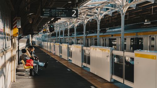 Free Commuters seated on a platform at a busy Tokyo train station under warm daylight. Stock Photo