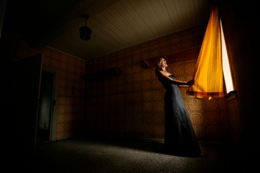 A woman in a dark dress opens a sunlit curtain in a vintage room, creating dramatic shadows.