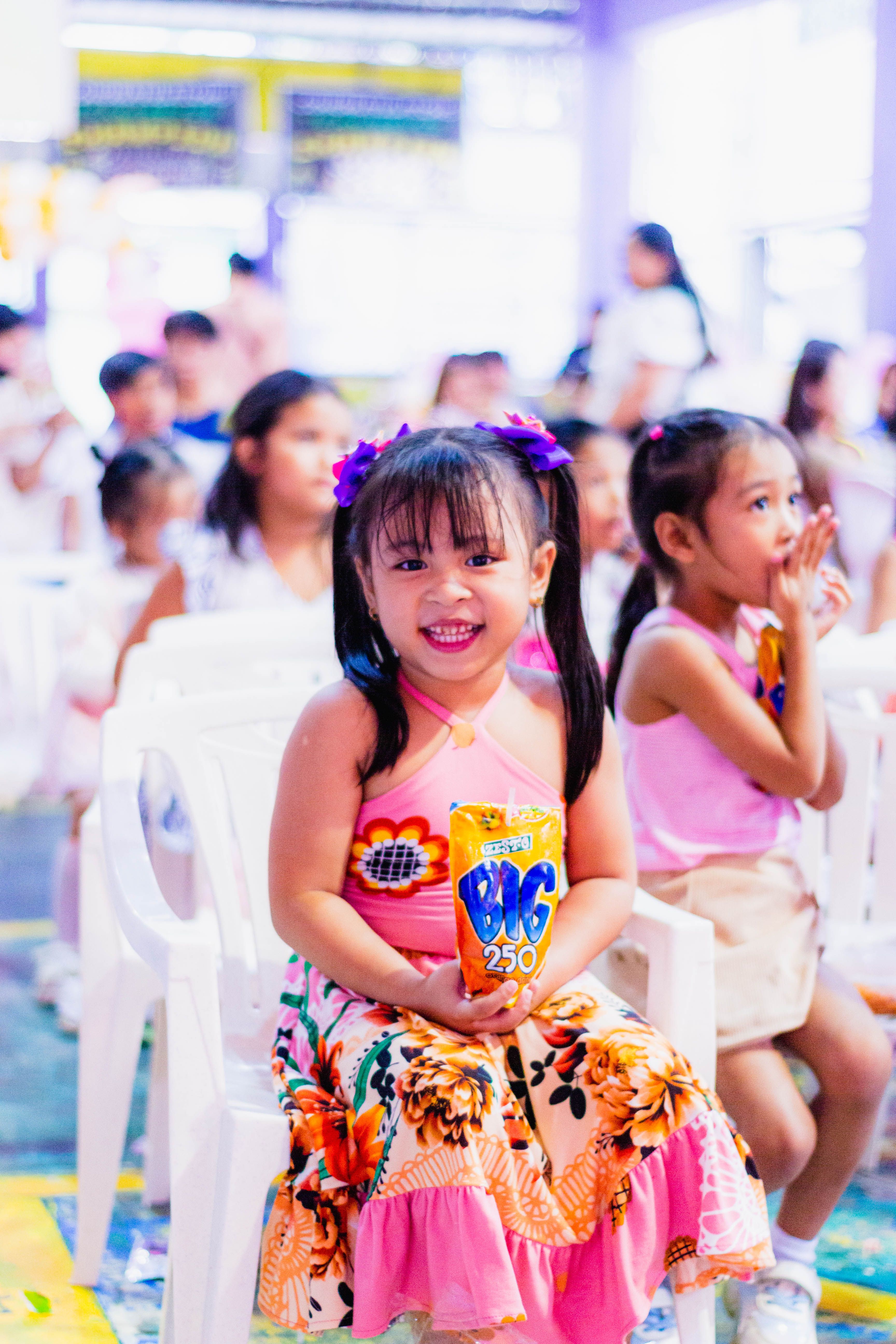 Children sitting indoors at an event, smiling and having fun, with a focus on bright colors and joyful expressions.