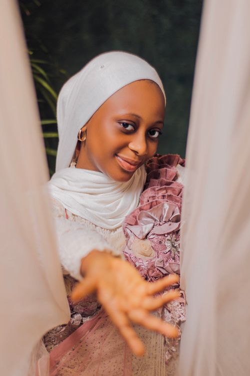 Free Smiling girl in a floral dress and hijab reaching out playfully indoors. Stock Photo