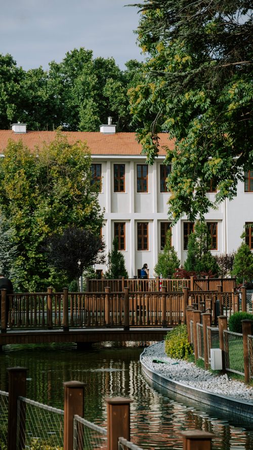 Free Serene view of an elegant building surrounded by lush greenery and a peaceful pond in a park setting. Stock Photo