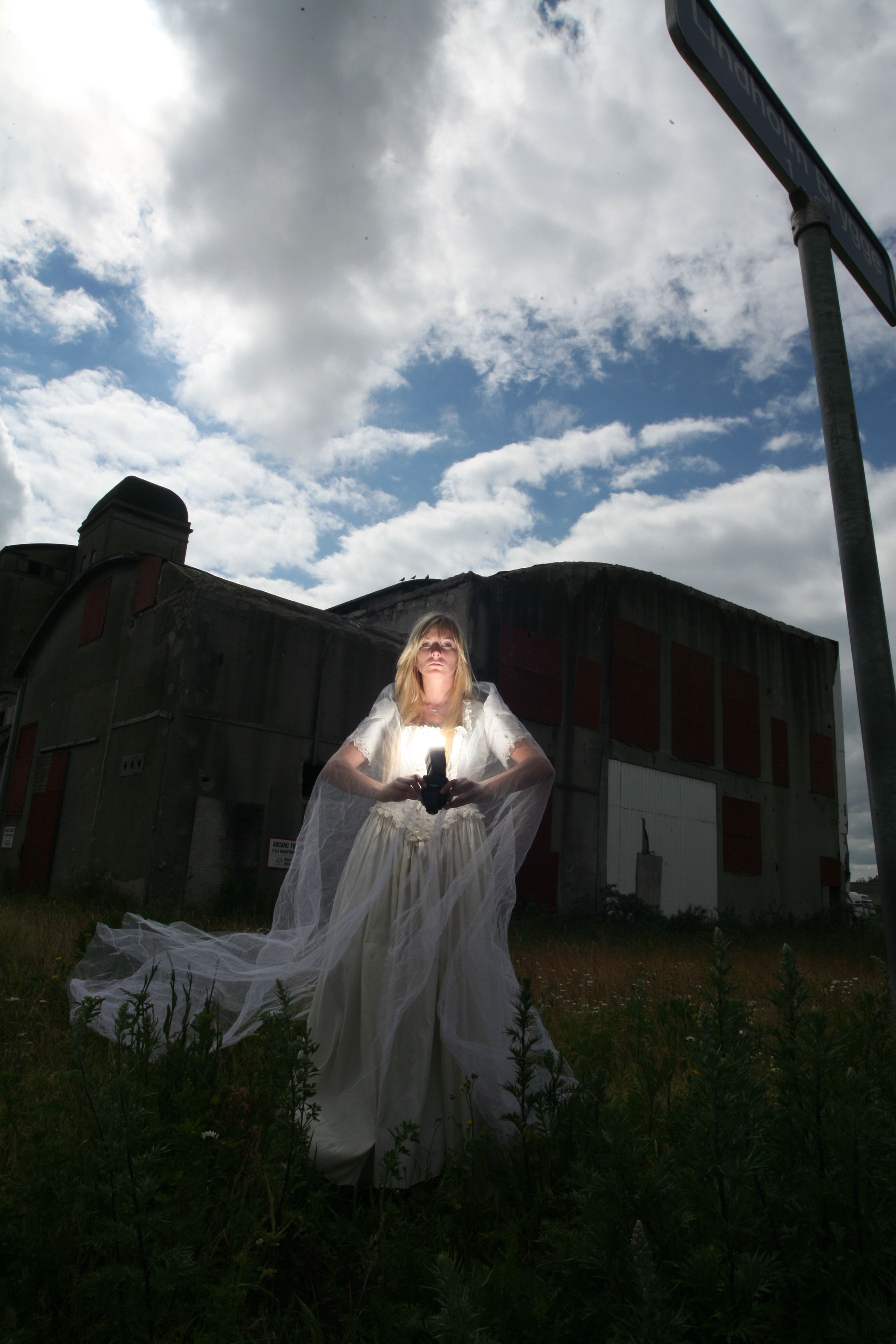 A woman in a wedding gown holding a flashlight under ominous clouds outdoors.