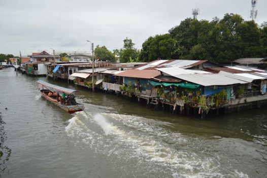 A scenic view of a traditional wooden boat navigating a canal beside rustic waterfront houses in Thailand.