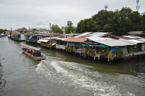 Free A scenic view of a traditional wooden boat navigating a canal beside rustic waterfront houses in Thailand. Stock Photo