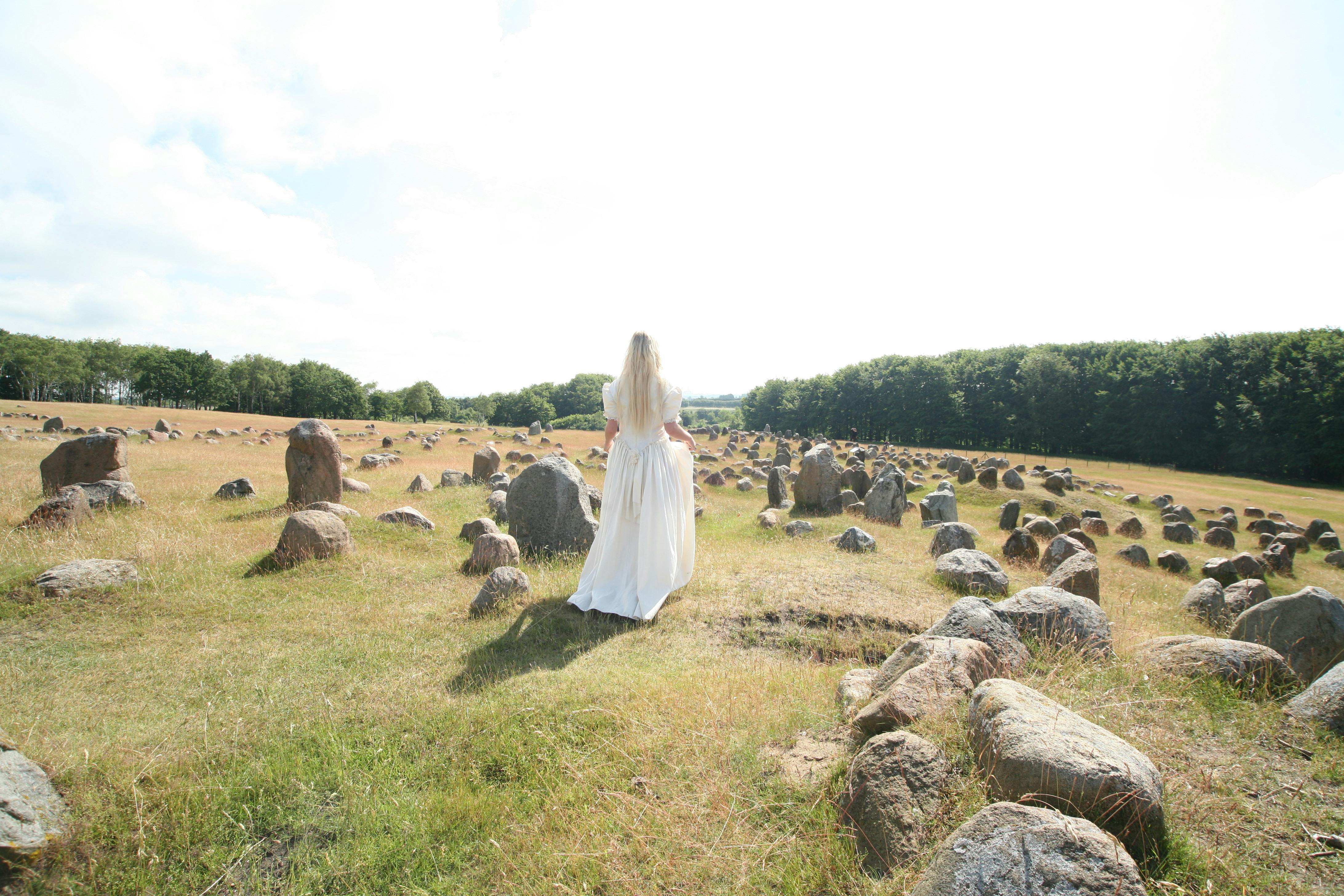 [ColoSach]-a-woman-in-a-white-dress-walks-through-a-historic-field-of-stones,-surrounded-by-nature.