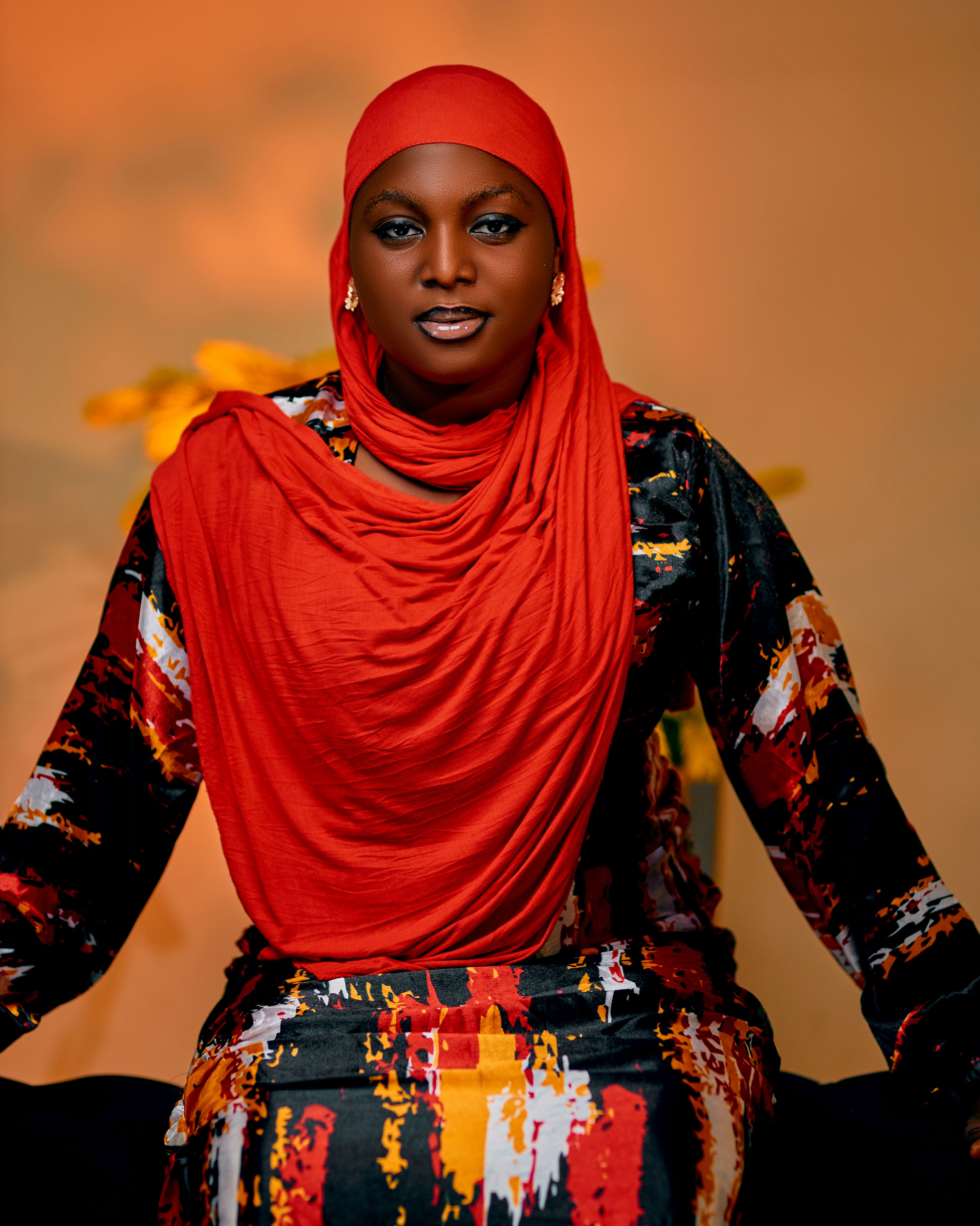 Portrait of an elegant woman wearing a vibrant red hijab with a colorful dress, set against a warm background.