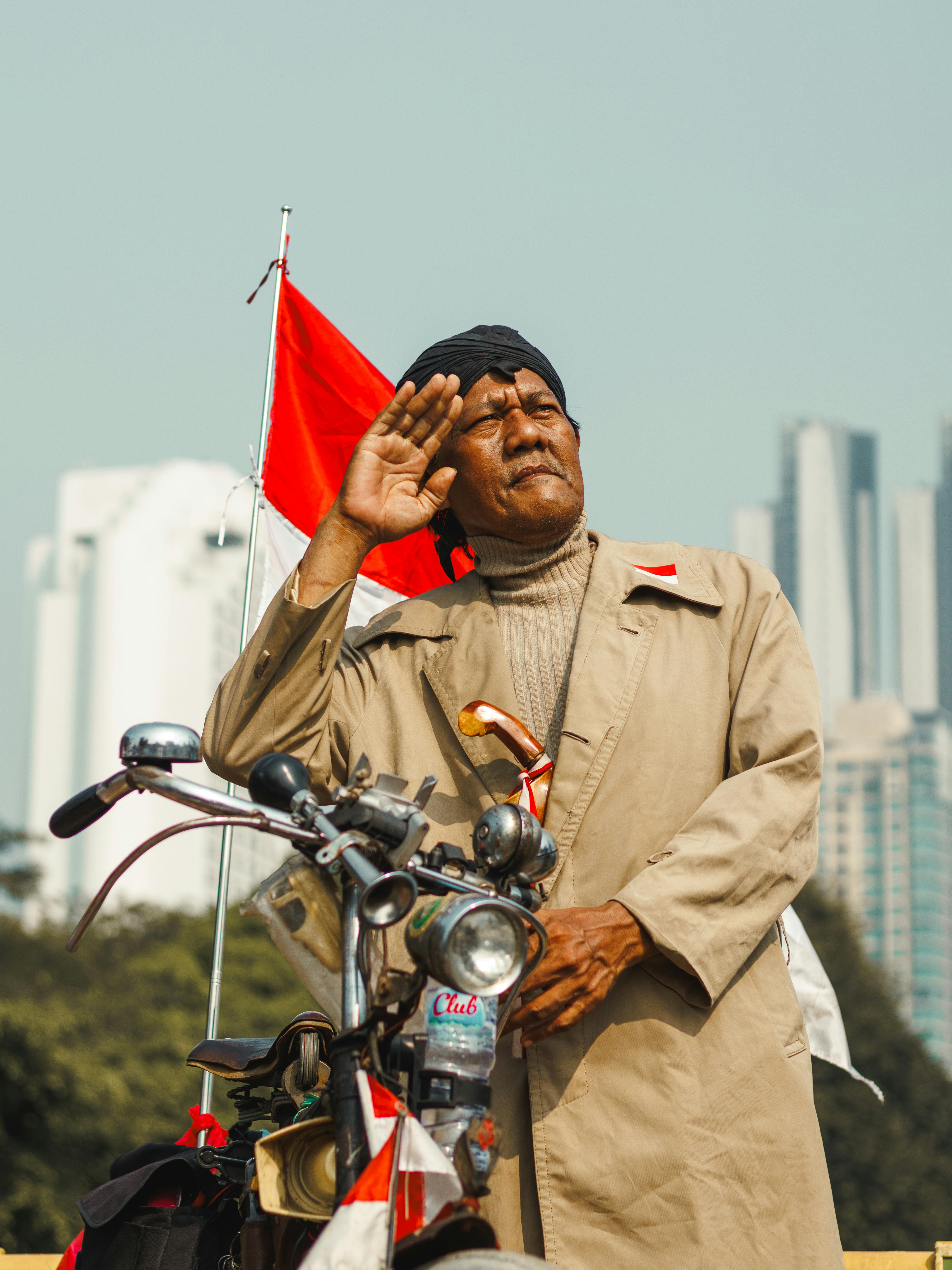 Elderly man on motorbike saluting with city skyline and red flag background.