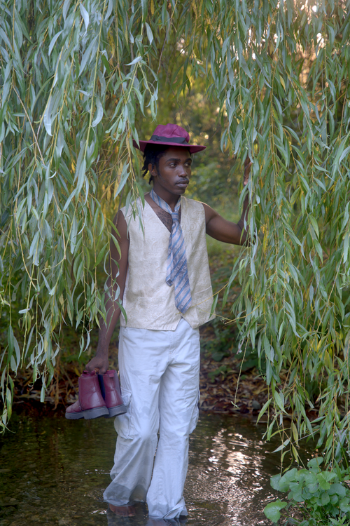 Free Man in nature wearing red hat, holding boots, surrounded by willow trees in a serene setting. Stock Photo