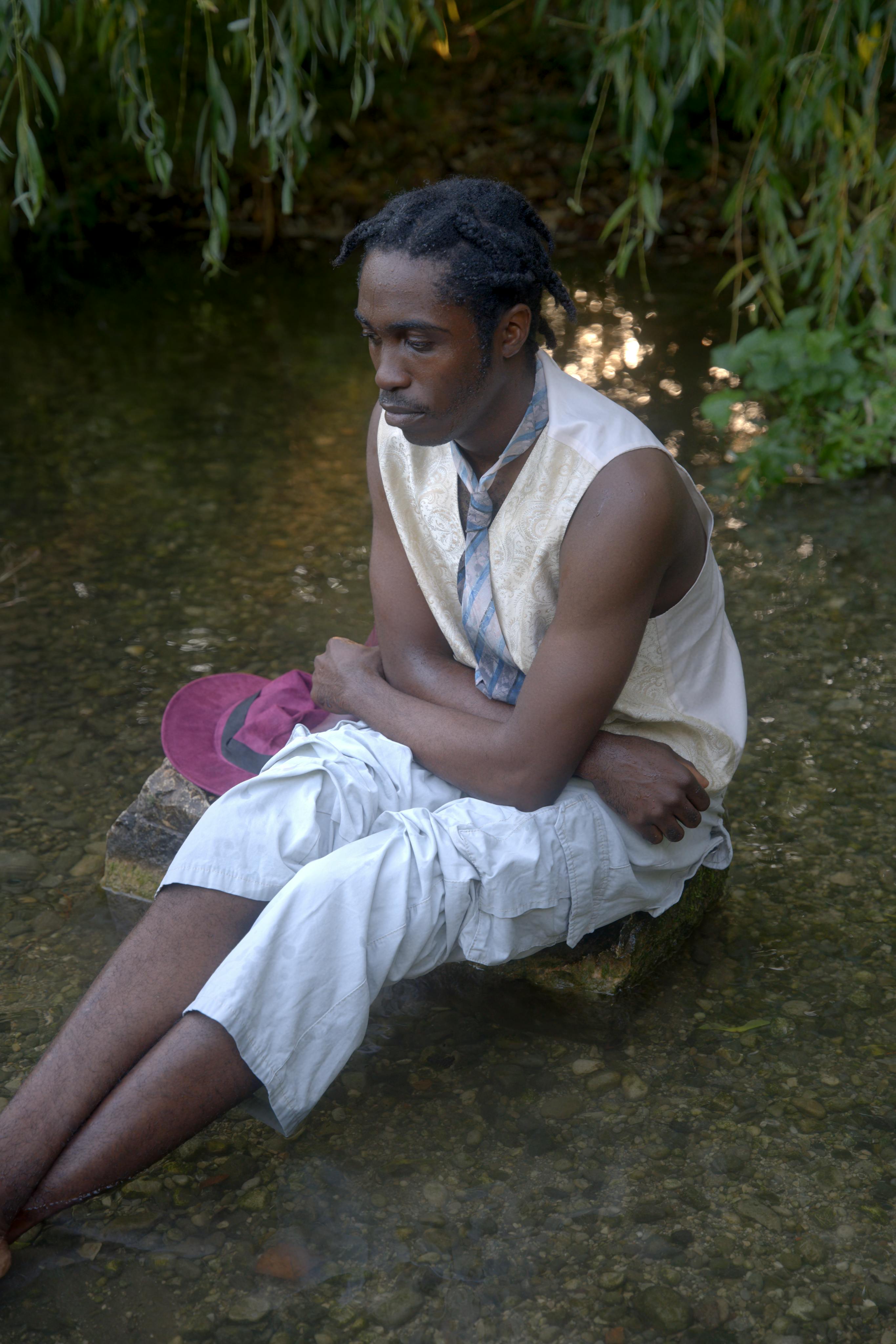 A thoughtful man sits on a rock in a serene forest stream surrounded by nature.