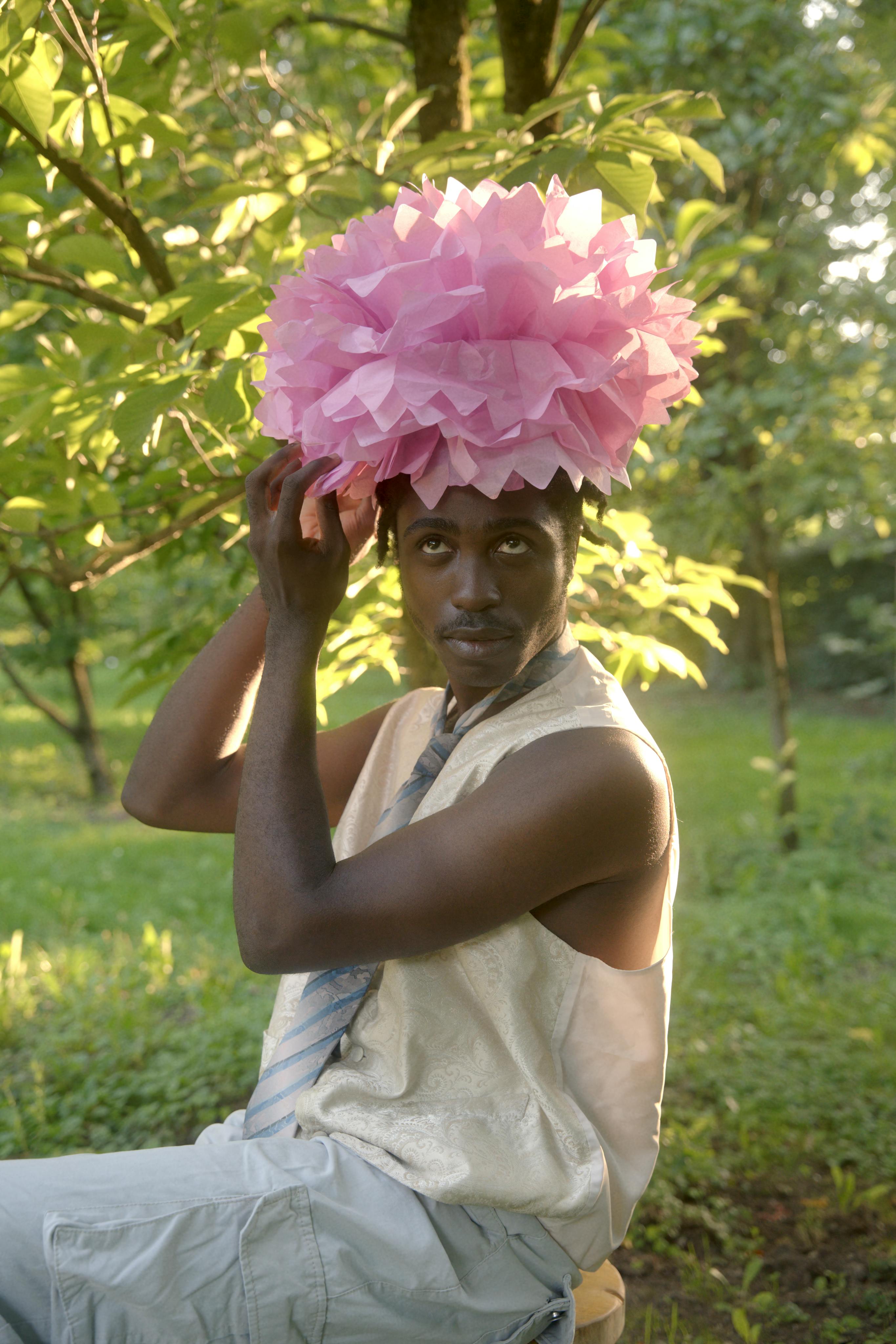 Portrait of a man wearing a pink paper flower hat, set in a sunny outdoor garden.