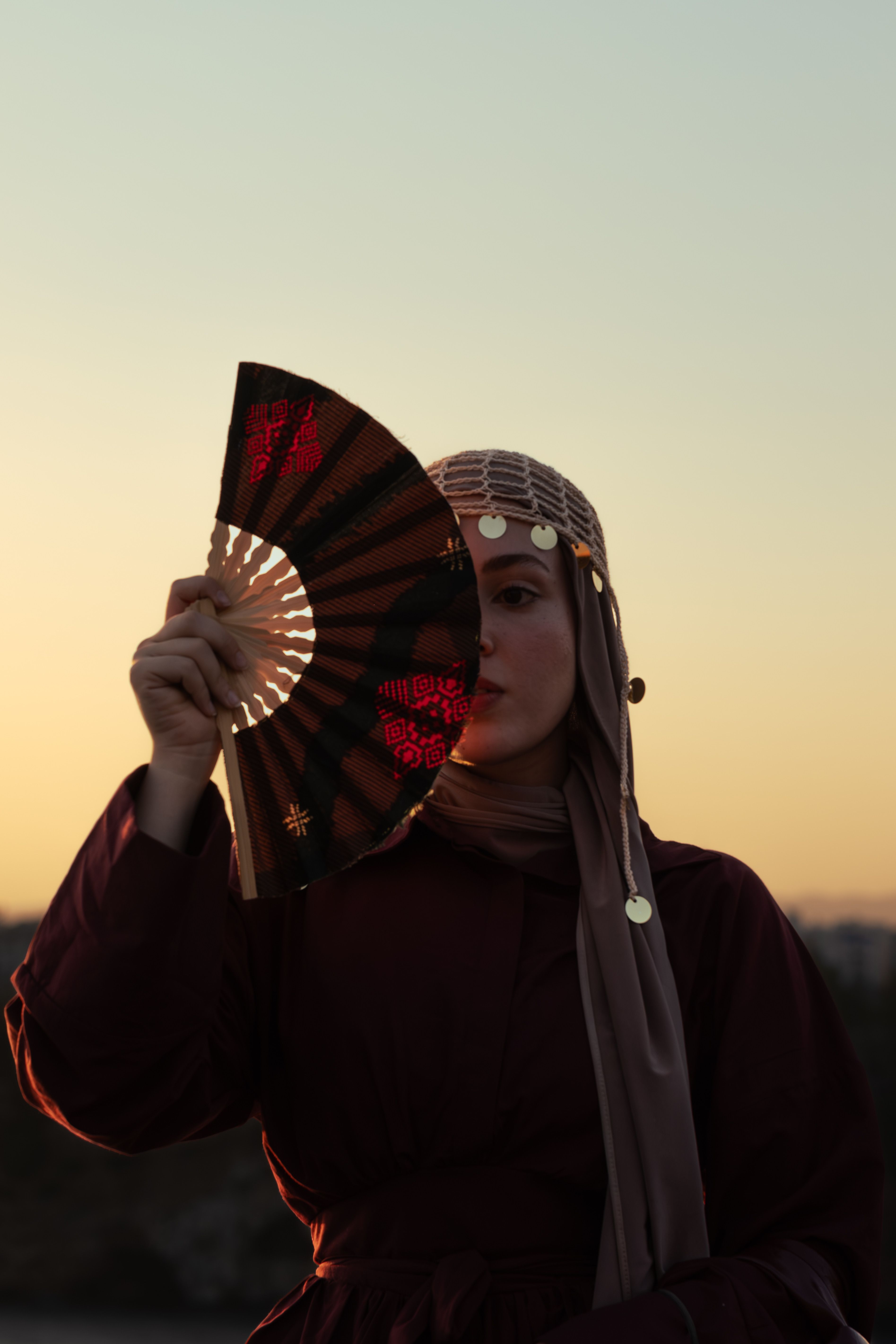 A woman in traditional attire holding a decorative fan at sunset, creating a mysterious silhouette.