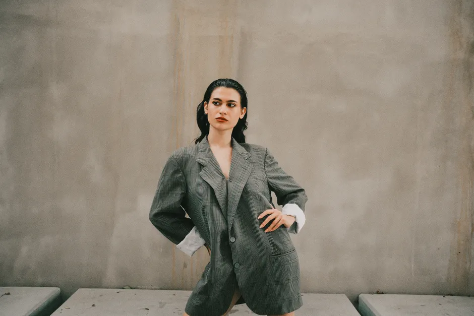 Femme en blazer oversize à carreaux posant devant un mur béton, style veste grande taille urbain