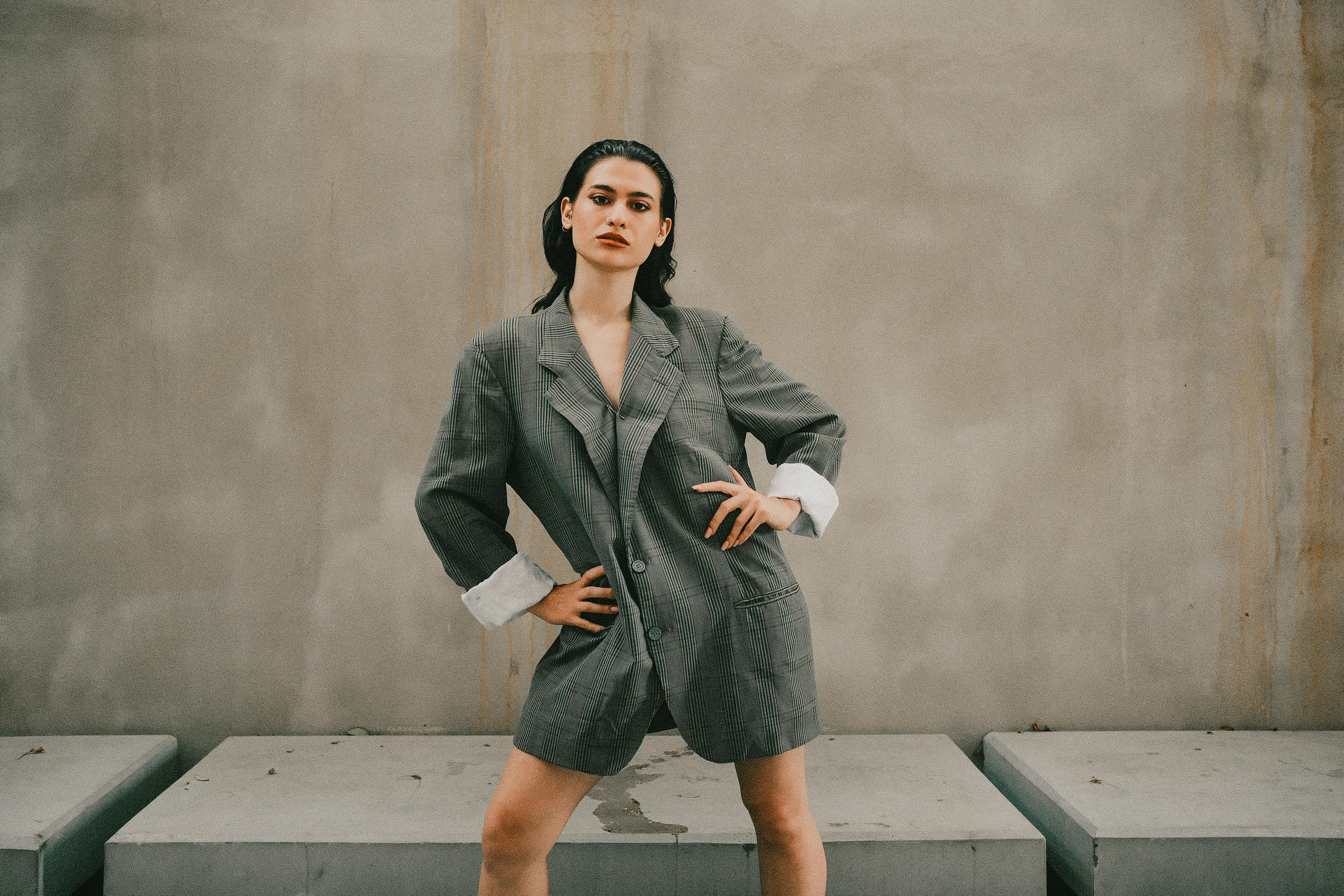 Stylish woman poses confidently in an oversized grey suit against a minimalist outdoor backdrop.