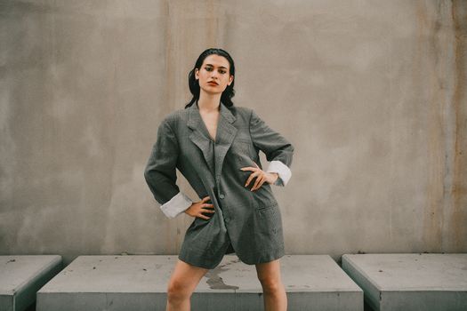 Stylish woman poses confidently in an oversized grey suit against a minimalist outdoor backdrop.