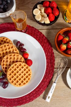 Top view of waffles with fresh strawberries, bananas, grapes, and maple syrup on a breakfast table.