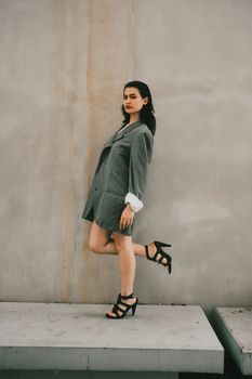Fashionable woman in gray blazer and heels posing against a minimalist concrete wall.