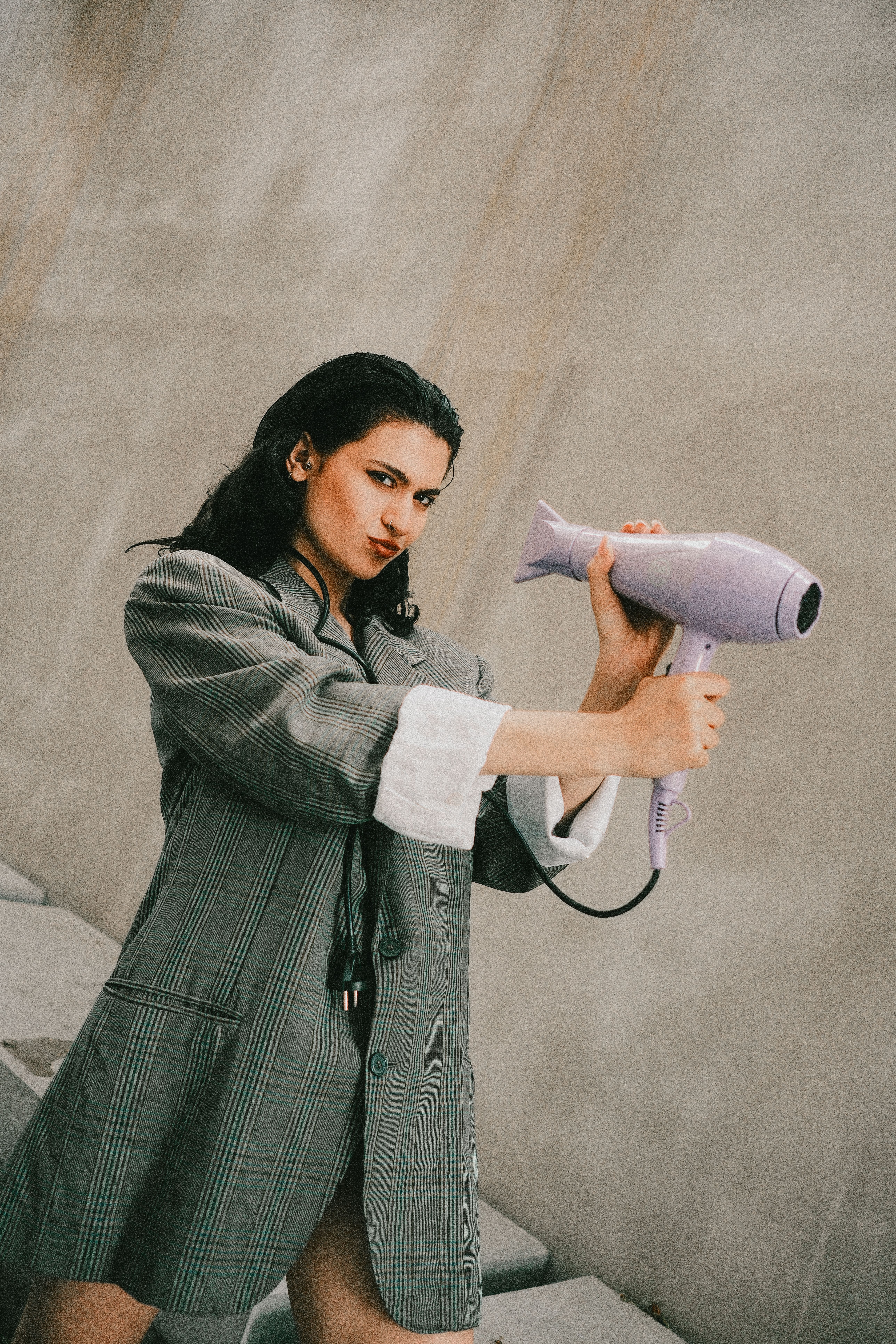 Stylish woman playfully posing with a hairdryer against a neutral background, showcasing fashion creativity.