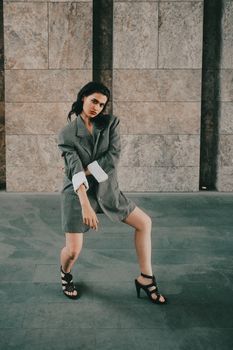 Fashionable woman poses confidently in oversized blazer and heels against an urban stone wall.