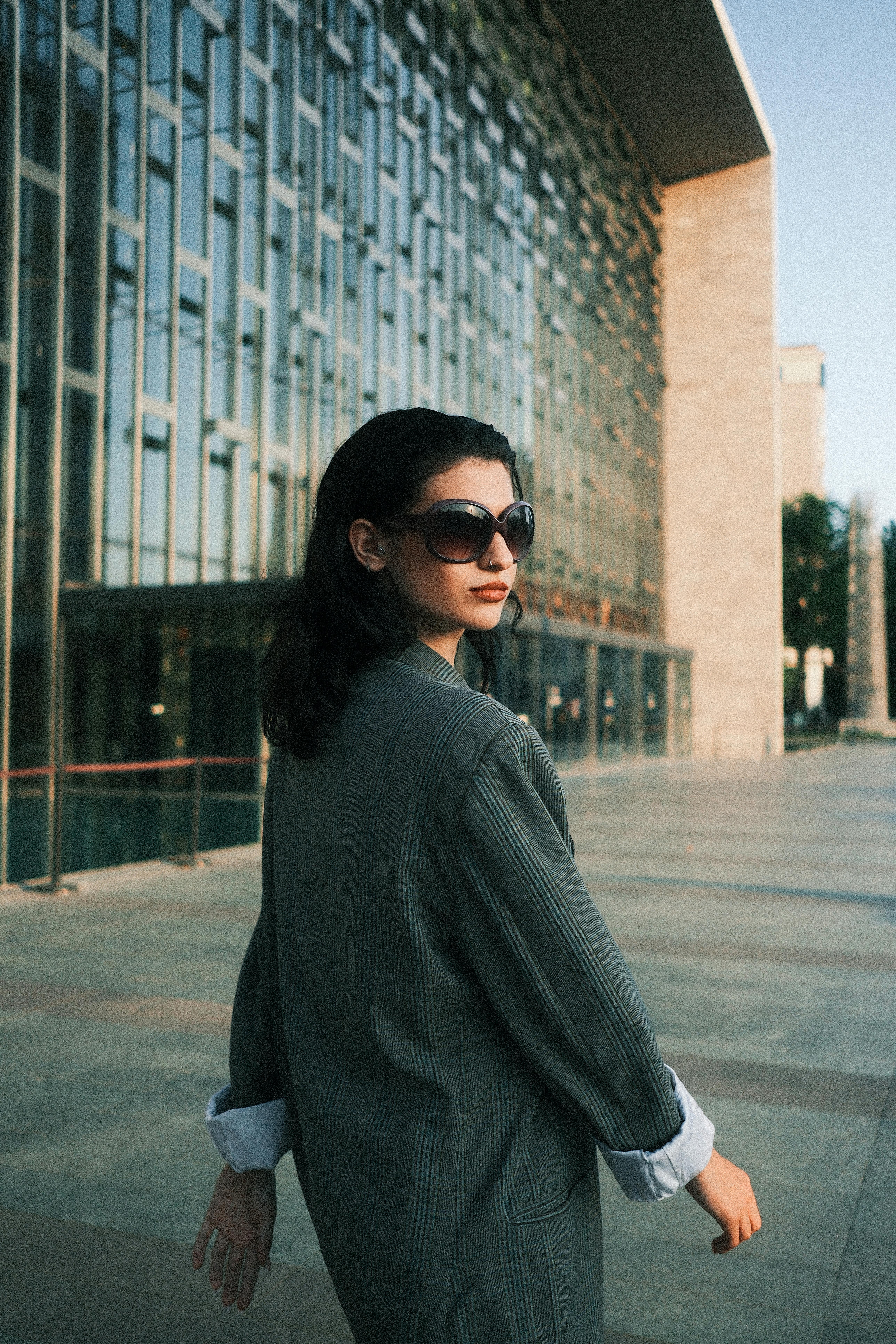 Stylish woman in sunglasses walks confidently past a sleek modern glass facade.
