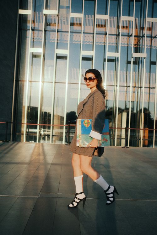 Free Stylish woman in a blazer carrying artwork outdoors beside a modern glass building. Stock Photo