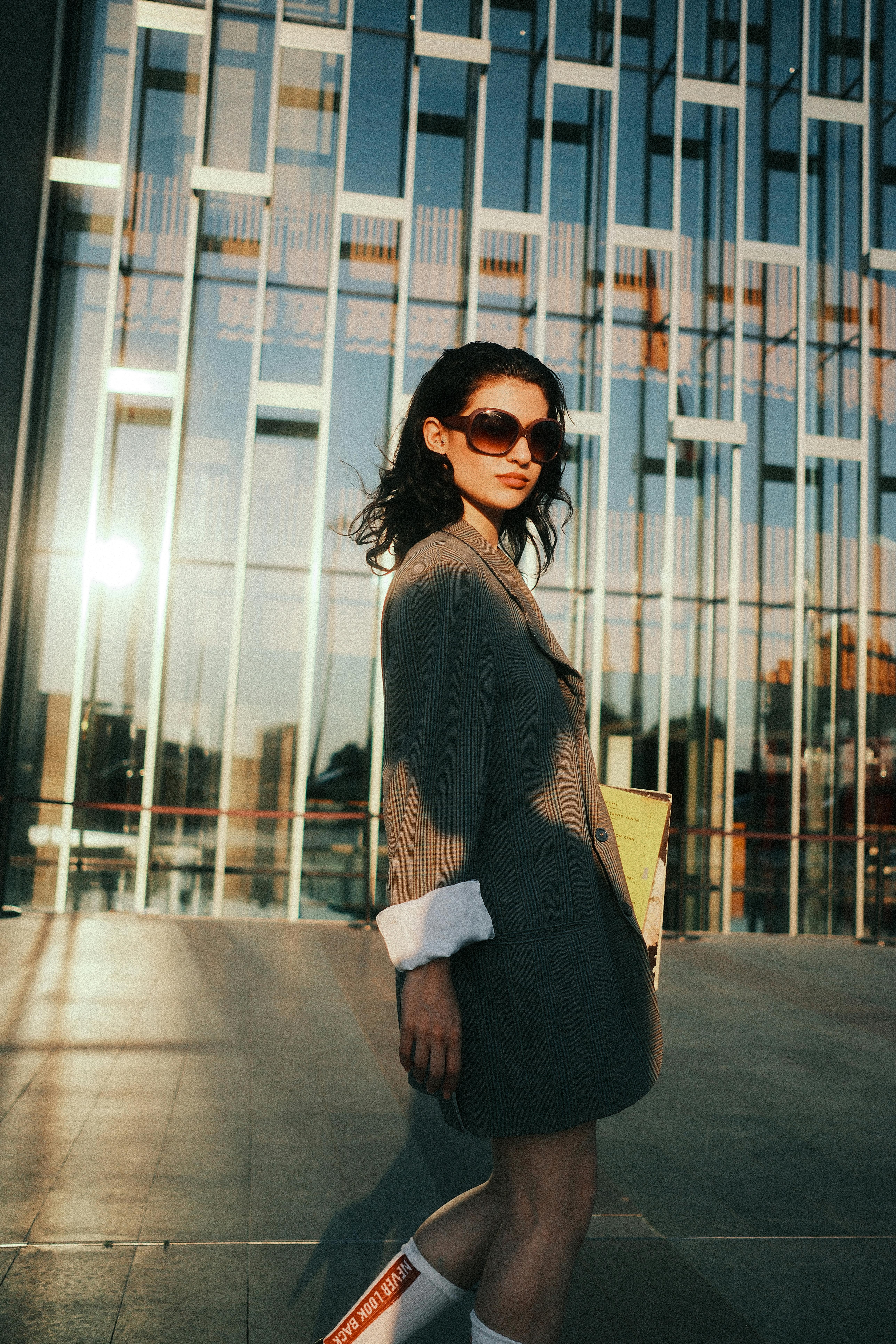 Stylish woman in sunglasses and blazer strolls by modern building facade at sunset.