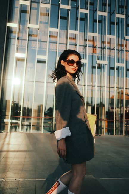 Stylish woman in sunglasses and blazer strolls by modern building facade at sunset.