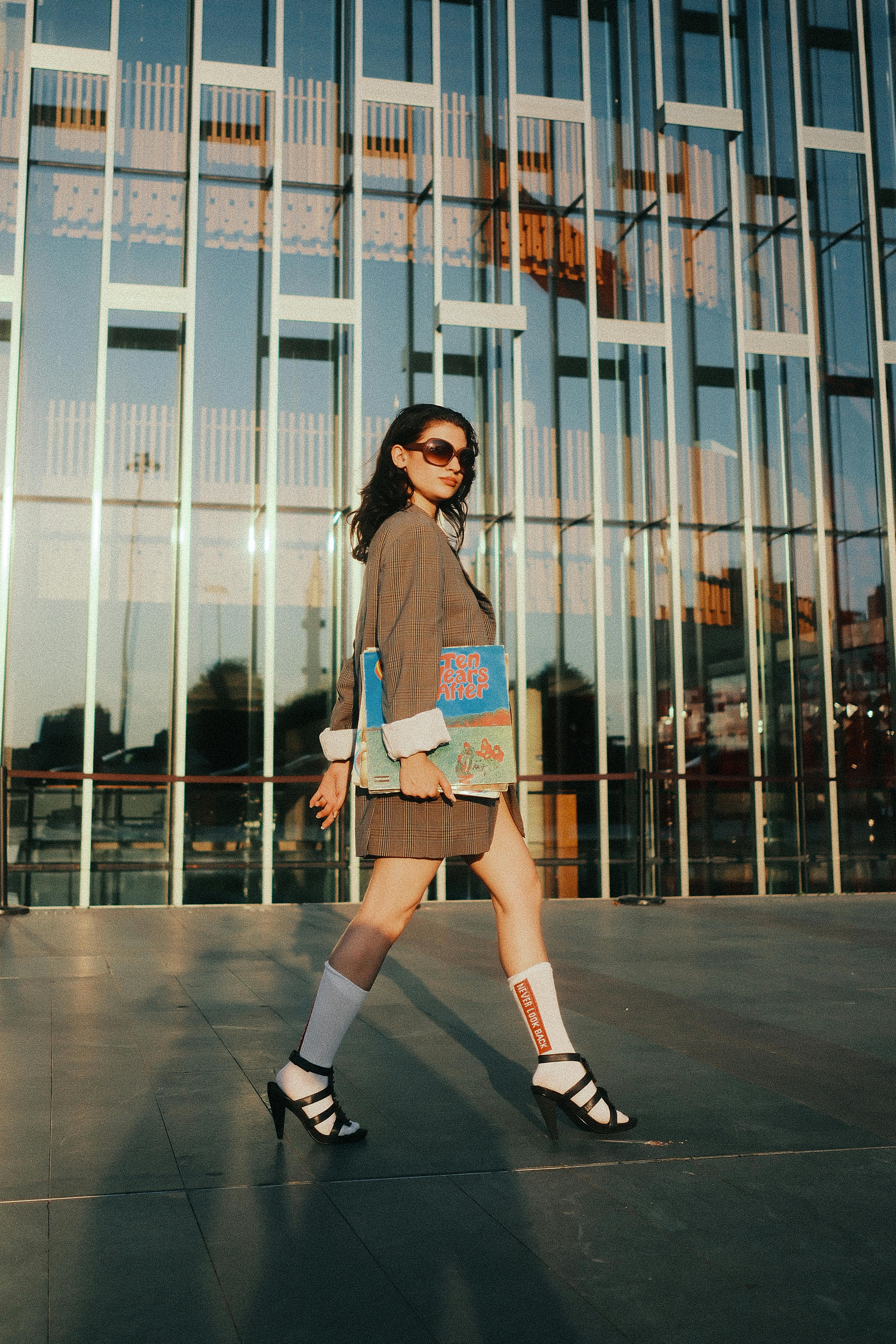 Fashionable woman in sunglasses and heels walking outside a glass building.