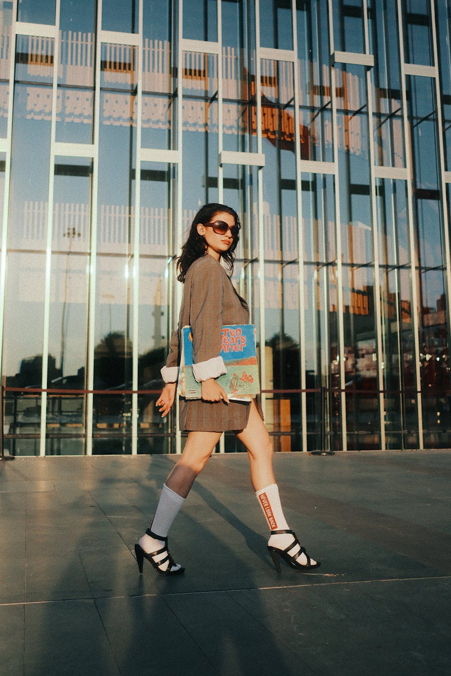 Fashionable woman in sunglasses and heels walking outside a glass building.