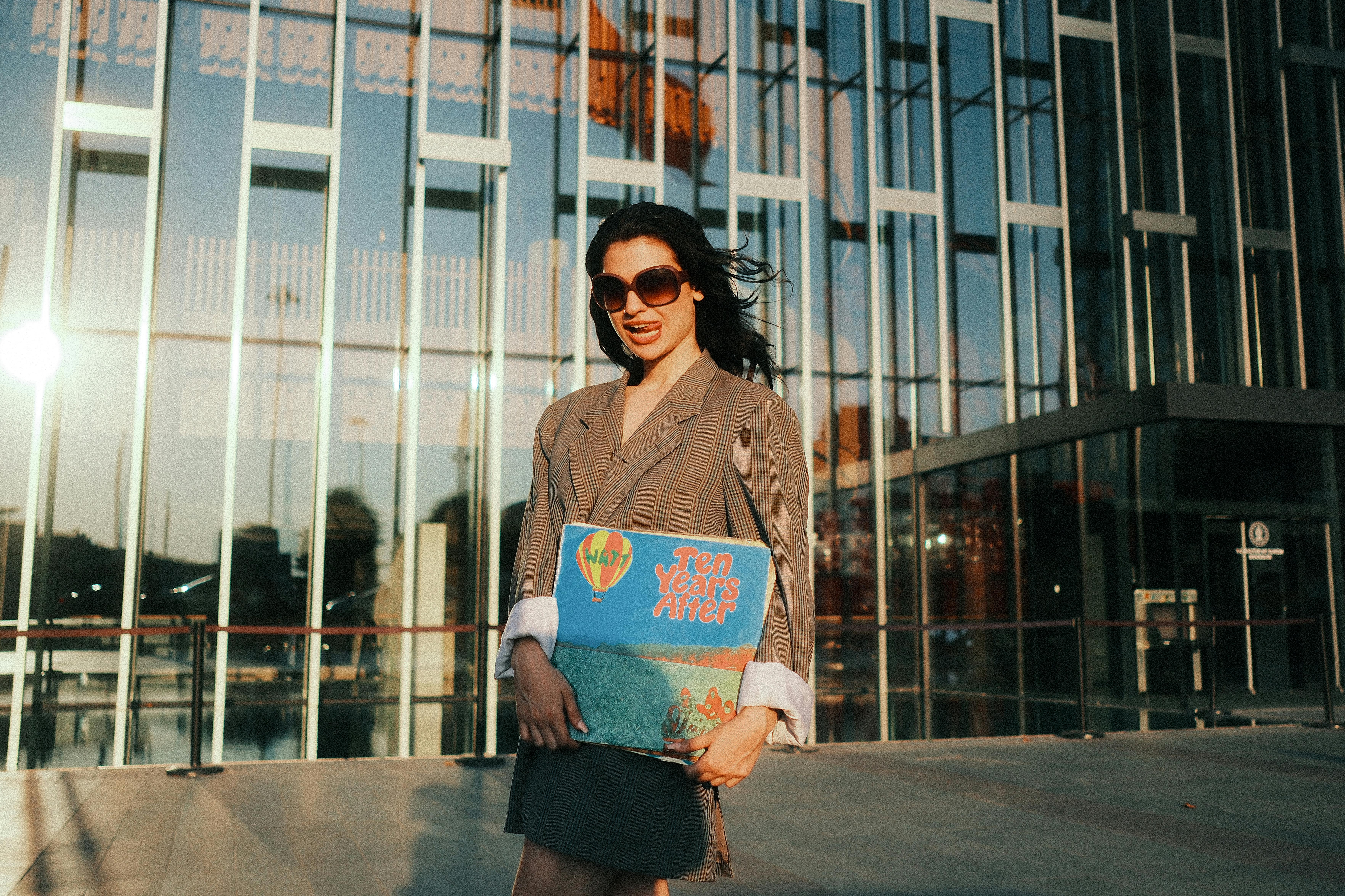 A fashionable woman holding a vintage album in front of a modern glass building during sunset.