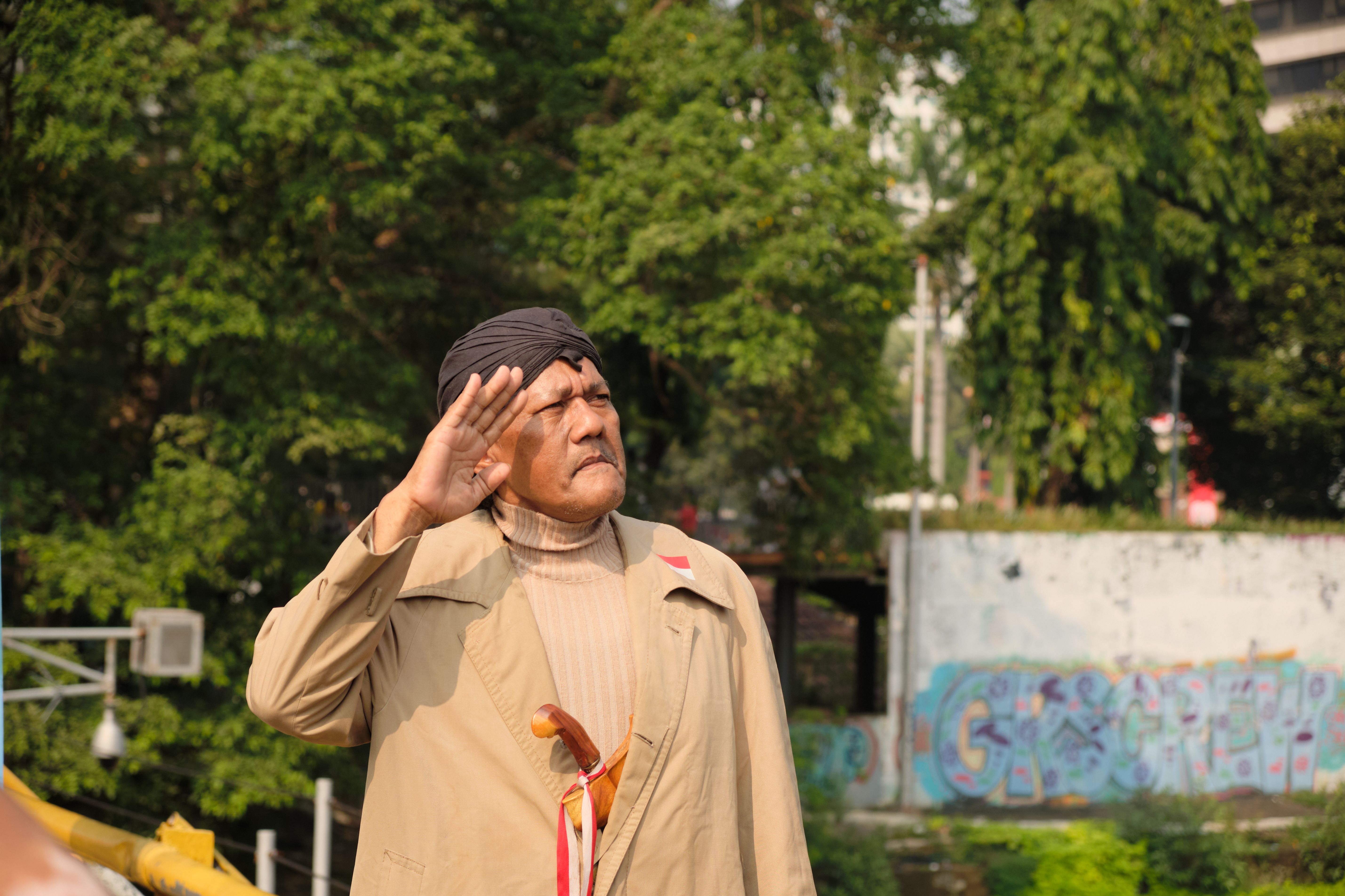 An elderly man salutes, embodying patriotism against a backdrop of greenery and urban graffiti.