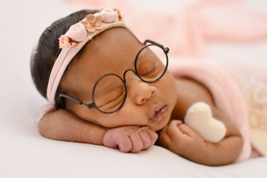 Cute baby with glasses and a floral headband sleeping peacefully, holding a small heart.