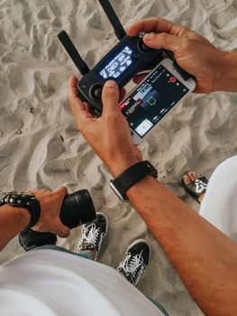Person operating a drone controller with smartphone display on a sandy beach setting.