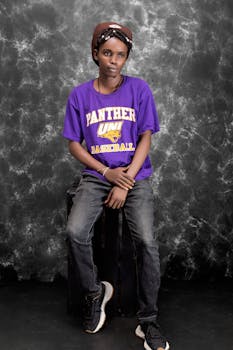 Young man in casual attire with a purple shirt, sitting on a stool against a textured gray background.