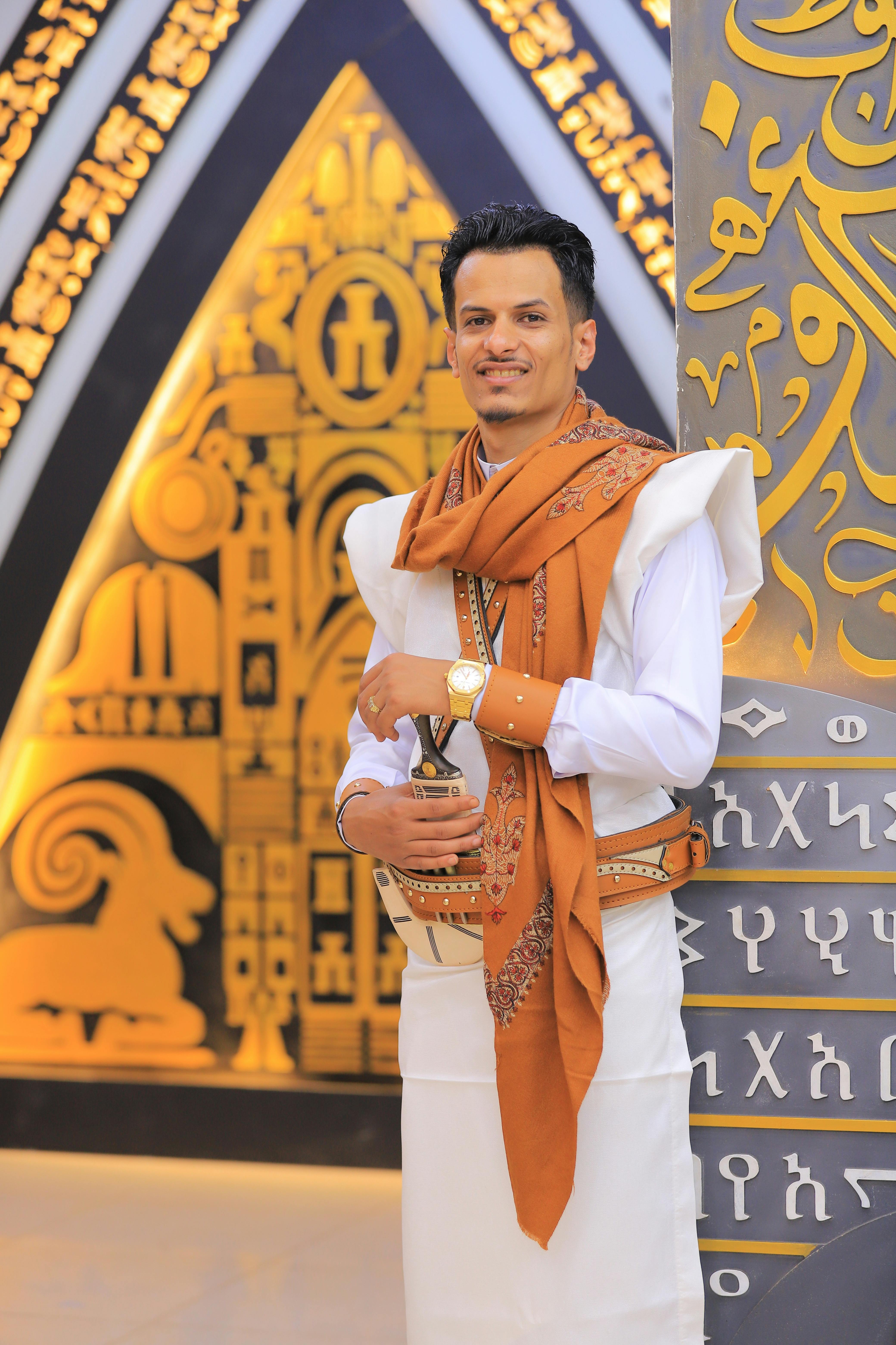 Young man in traditional attire stands against a decorative background with cultural motifs.