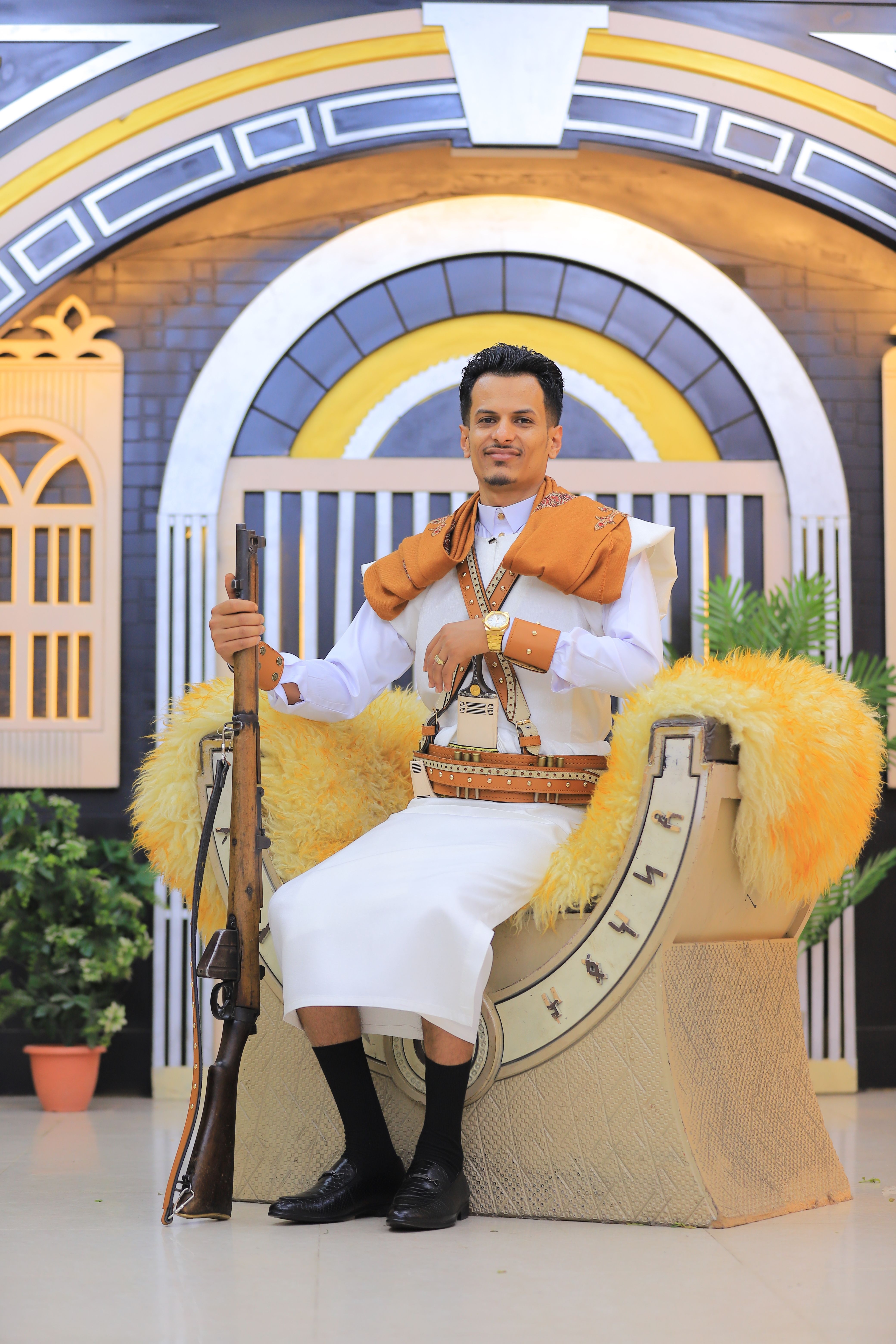 A man in traditional Middle Eastern attire seated with a rifle indoors.