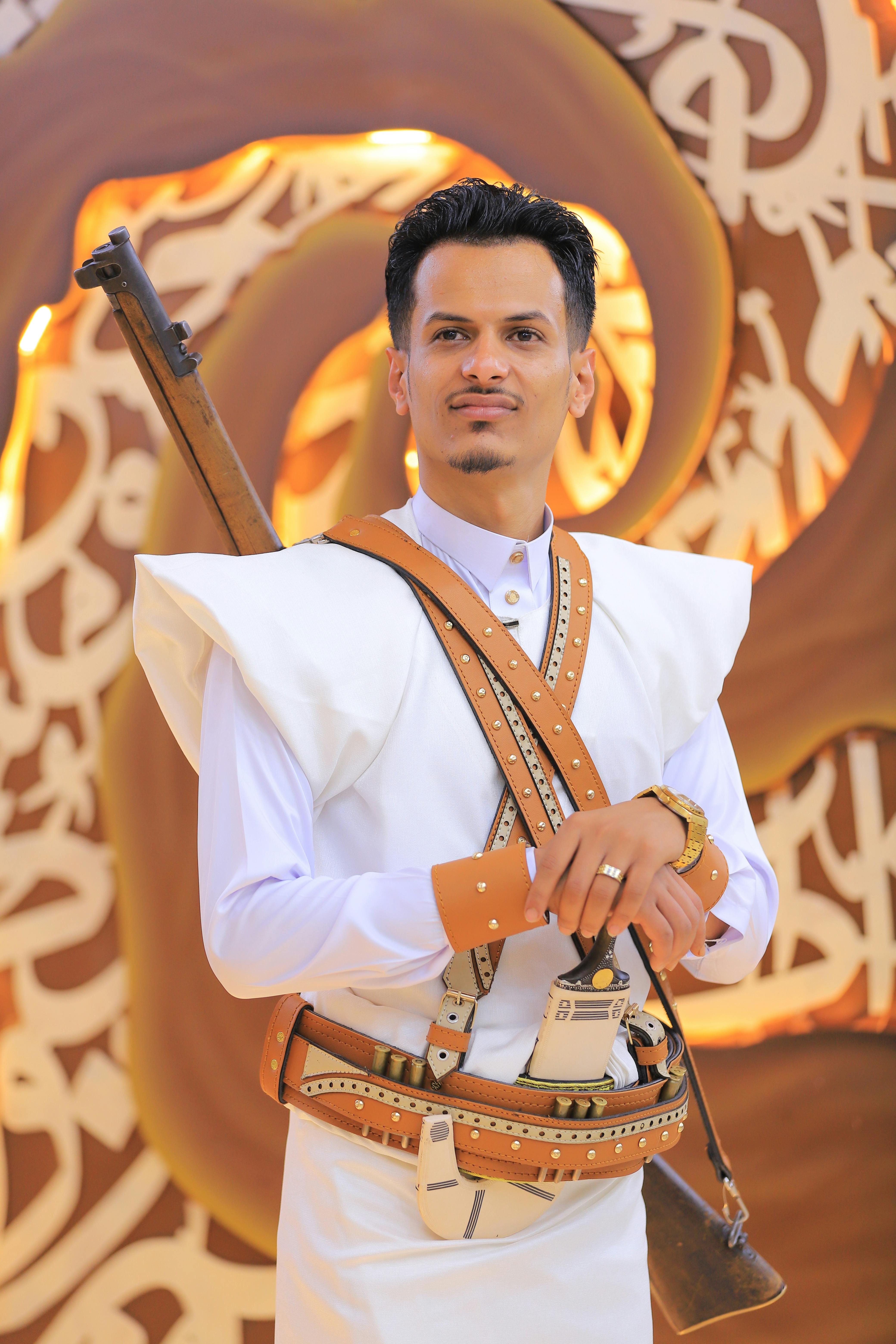 Man in traditional outfit with a rifle, against an ornate background with intricate patterns.