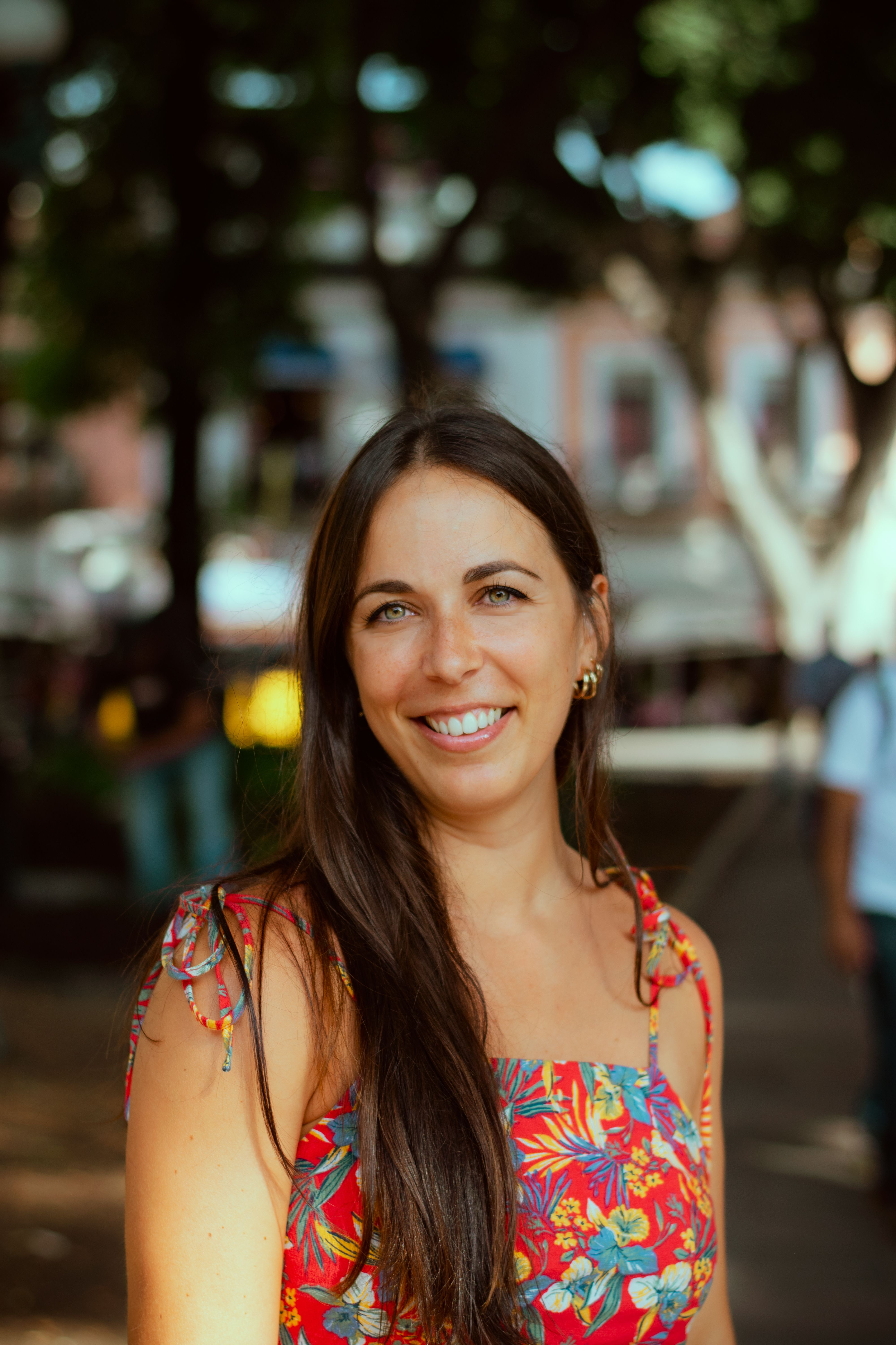 Portrait of a smiling woman in a vibrant floral dress, standing outdoors in a sunny park.