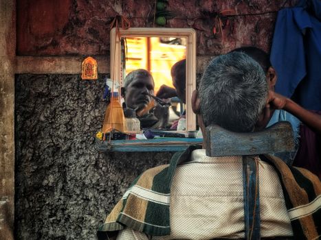 Rustic street barber setup with a mirror reflecting an elderly man's face during a shave.