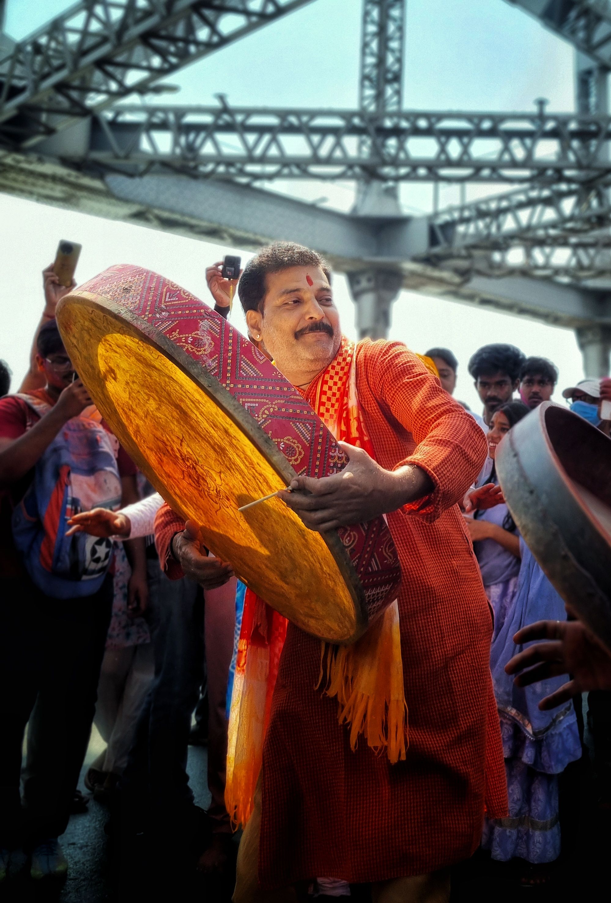Man energetically playing a traditional drum during a lively festival under a bridge.