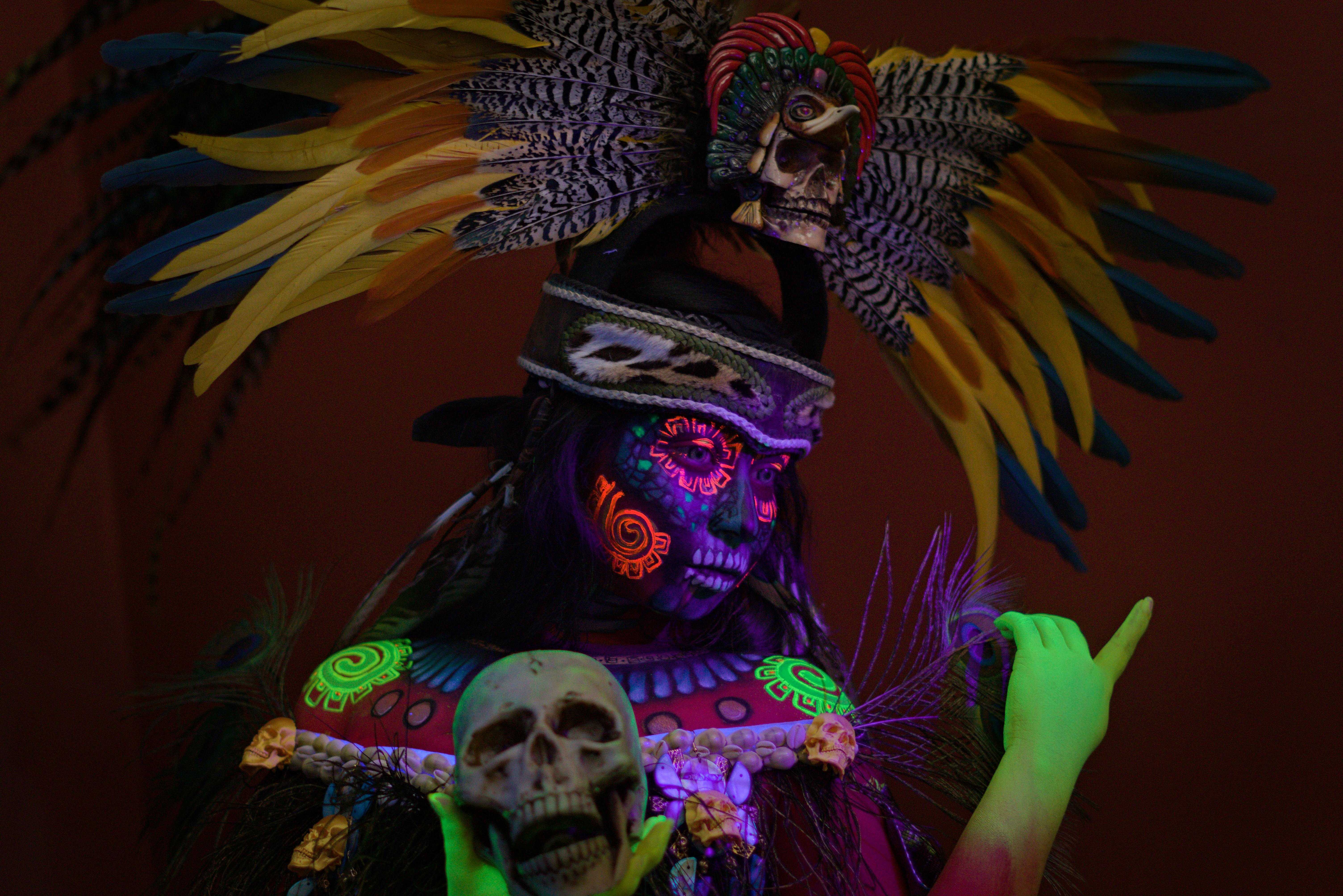Person in colorful traditional attire and painted face, holding a skull with vibrant lighting.